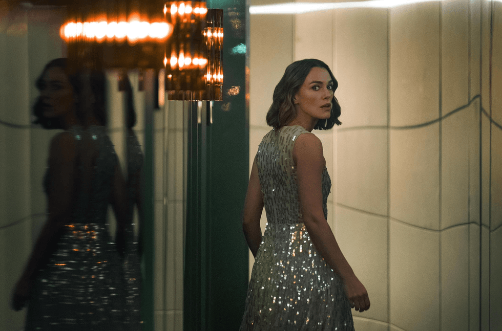 Woman in a shimmering dress looks back in a dimly lit, reflective room.