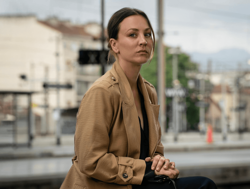Woman in a tan coat sits at a train station, gazing intently ahead.