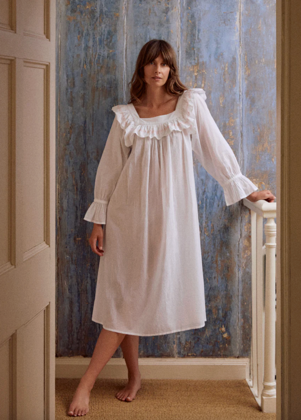 Woman in a white nightgown stands by a textured blue wall.