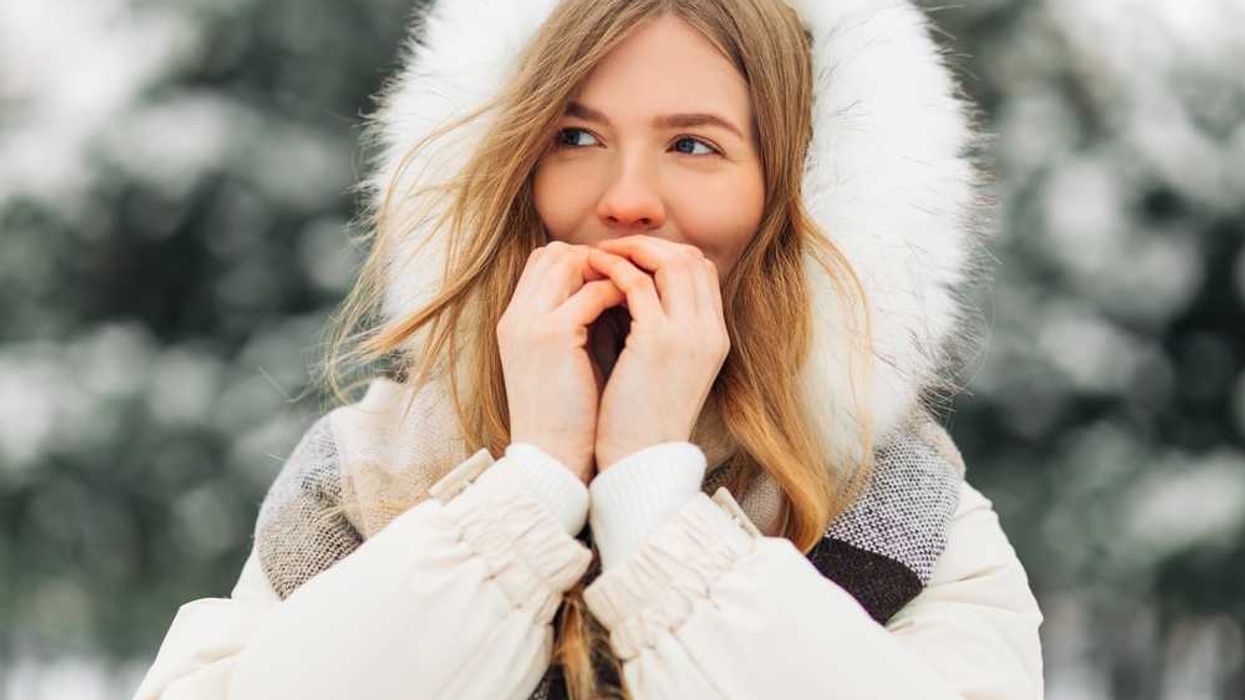 Woman in a winter coat and fur hood standing in snowy landscape, holding her hands to her mouth.
