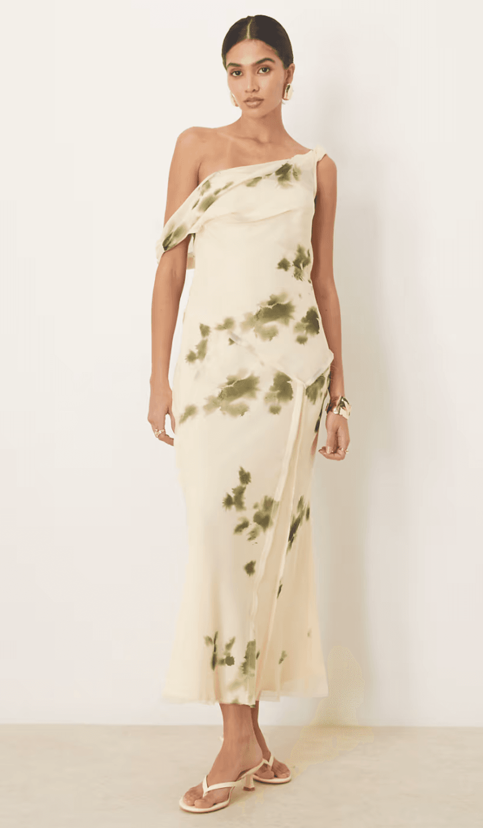 Woman in an off-shoulder cream dress with green floral pattern, standing indoors.