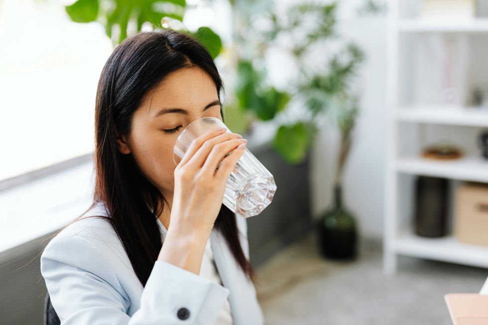 Woman in an office drinks water from a glass beside a leafy plant.
