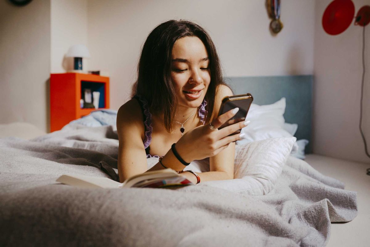 Woman in bed smiling at her phone, with an open book nearby.