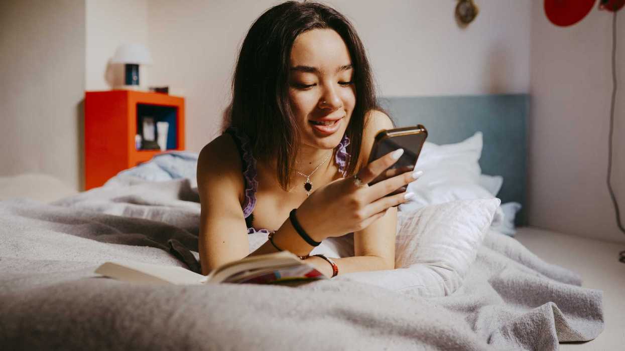 Woman in bed smiling at her phone, with an open book nearby.