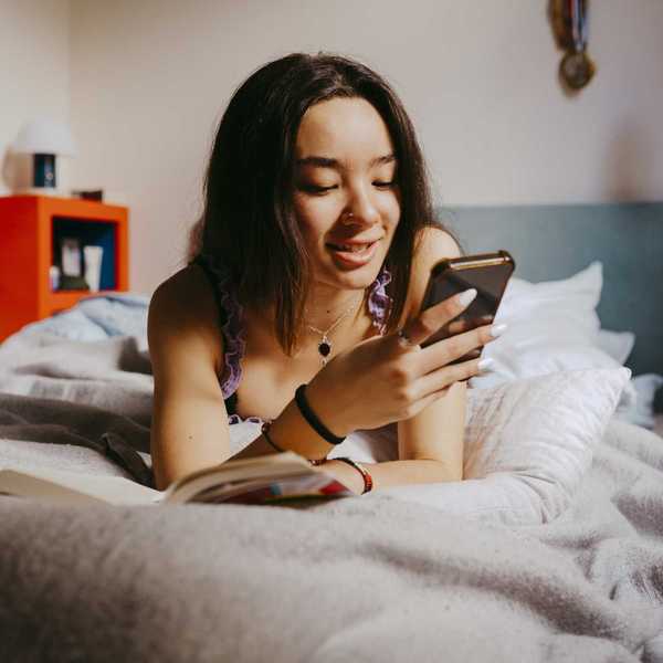 Woman in bed smiling at her phone, with an open book nearby.