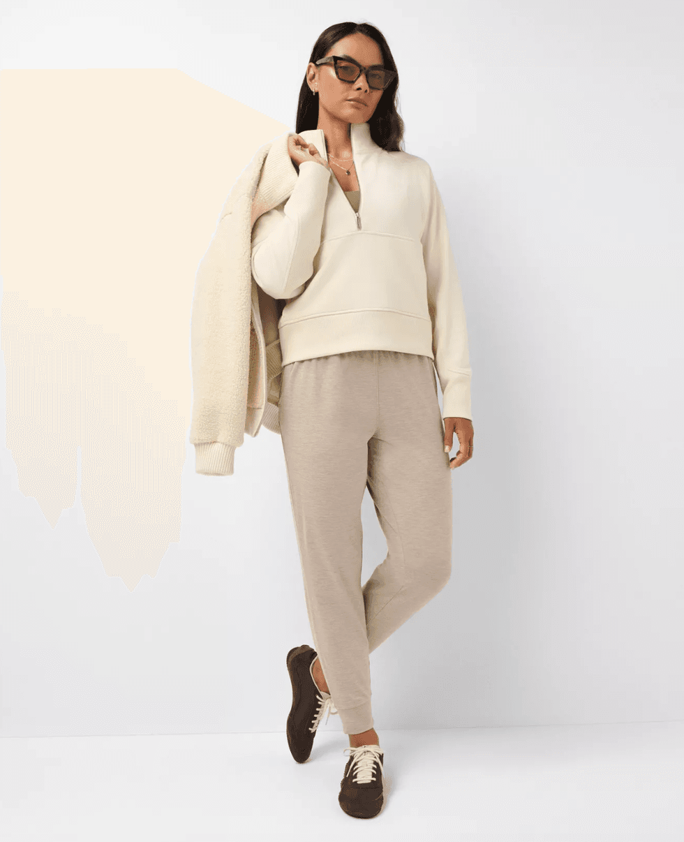 Woman in beige casual outfit with jacket and sunglasses, leaning against a wall.