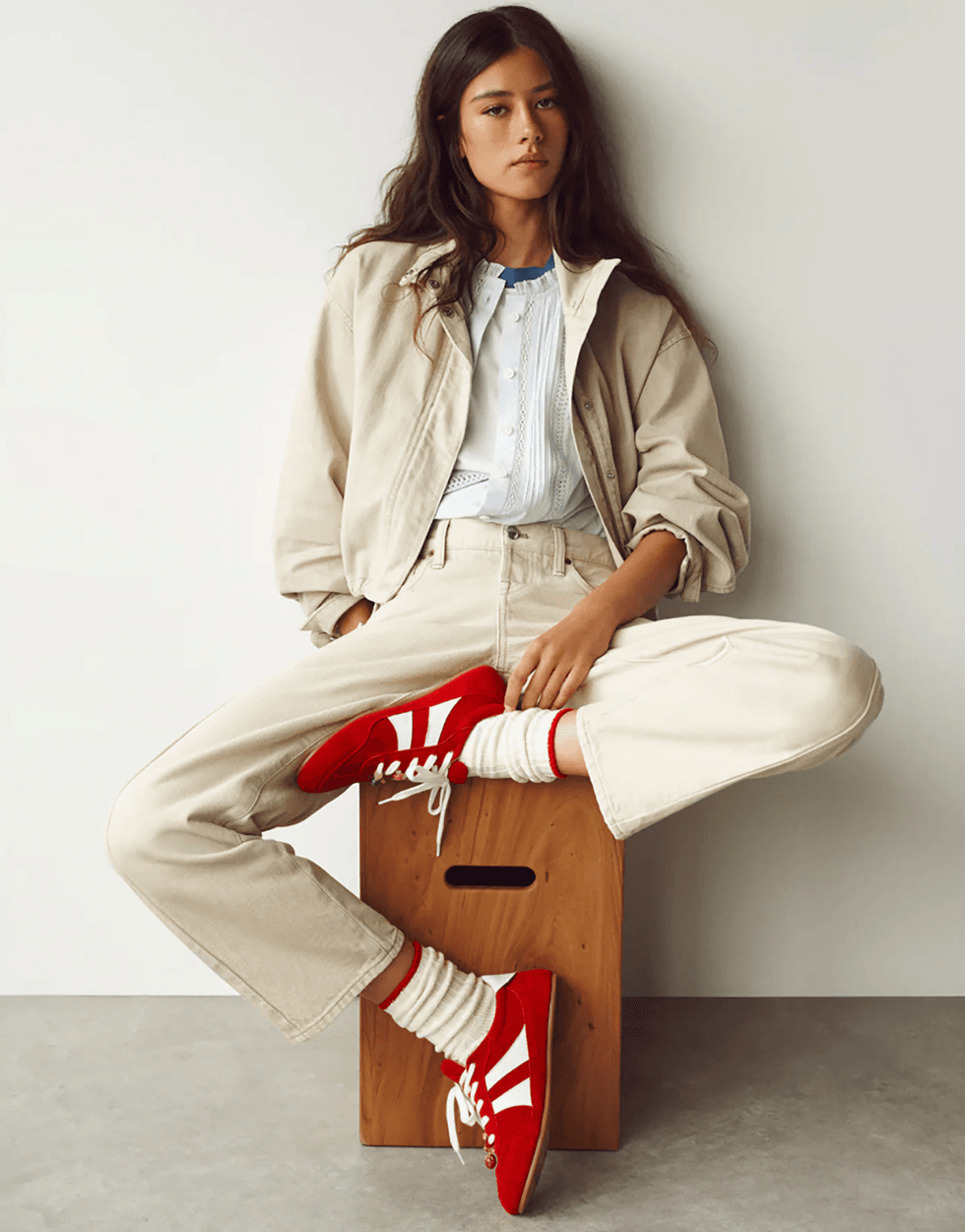 Woman in beige outfit and red shoes sits on wooden box against white background.