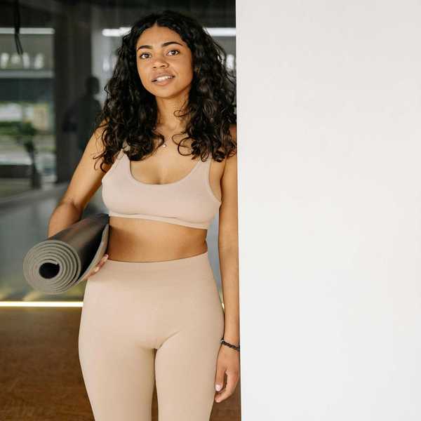 Woman in beige workout wear holding a yoga mat, standing by a bright wall.