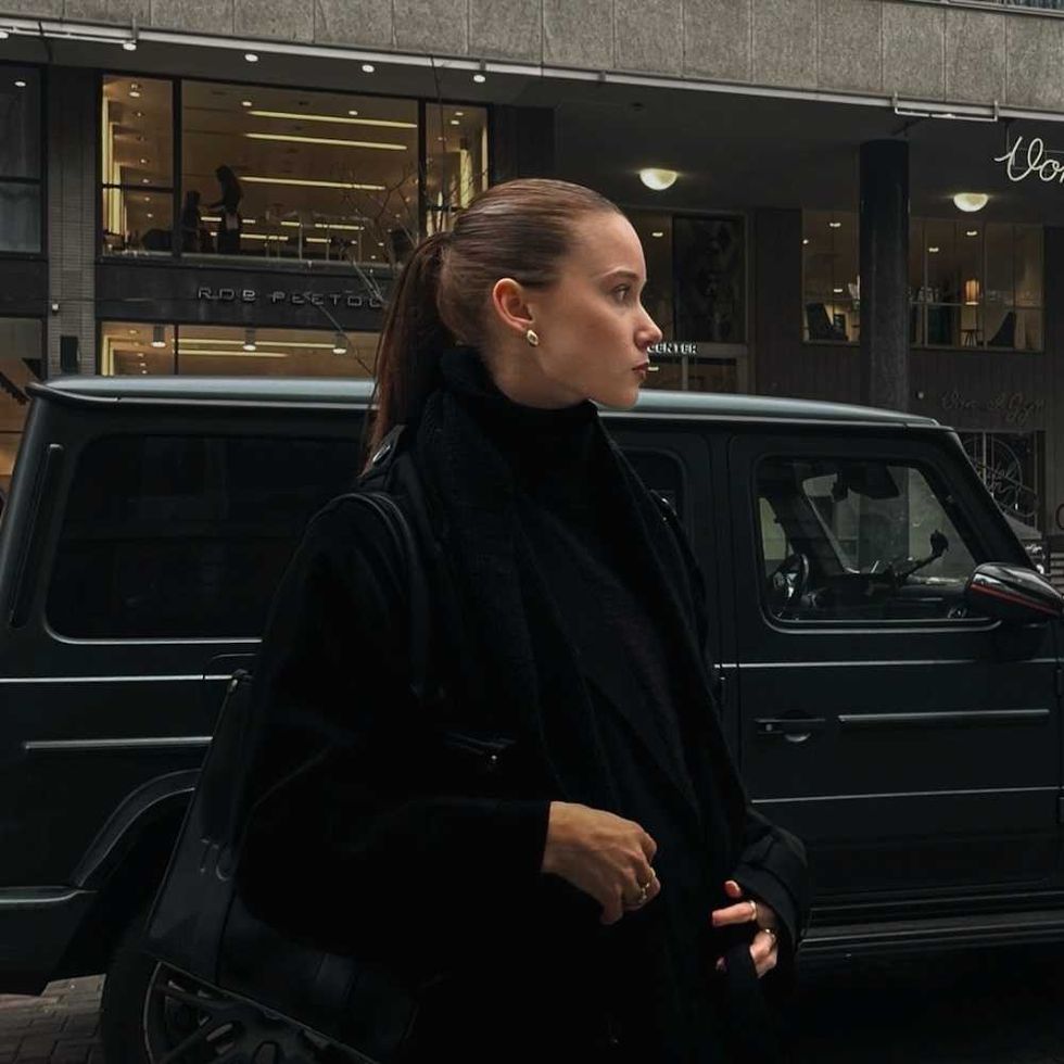 Woman in black coat stands by dark SUV in urban scene.