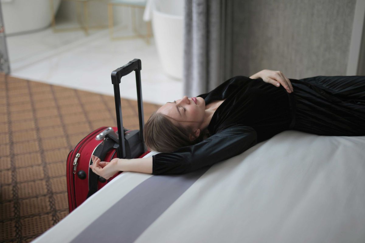 Woman in black dress relaxing on a bed with a red suitcase nearby.