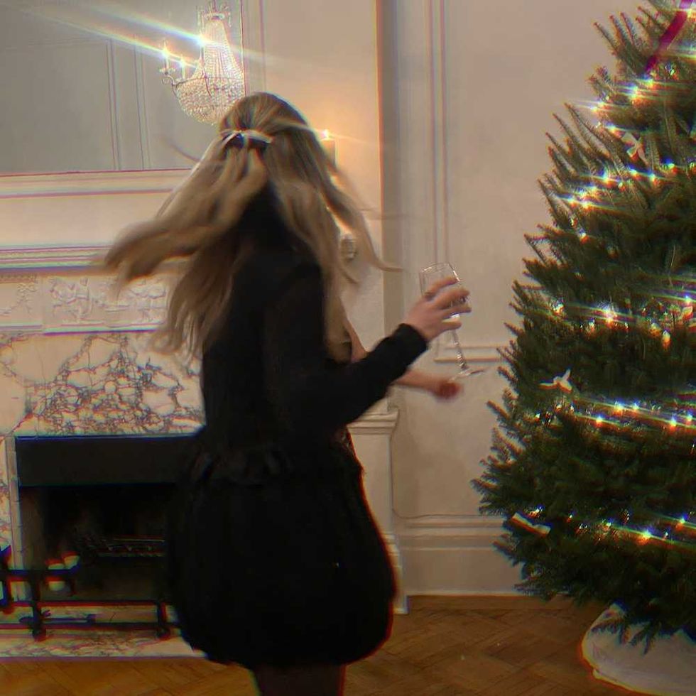 Woman in black dress twirls with drink near decorated Christmas tree indoors.