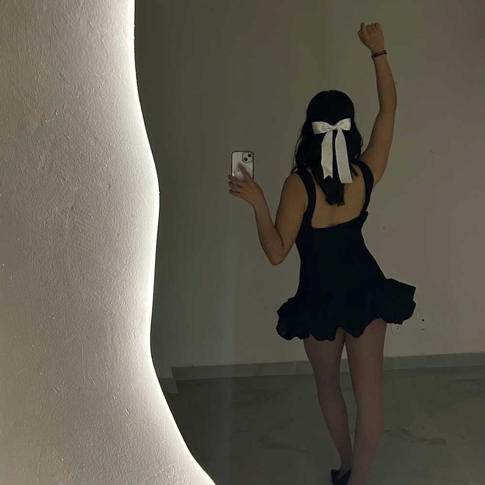 Woman in black dress, white bow, takes mirror selfie in dim room.