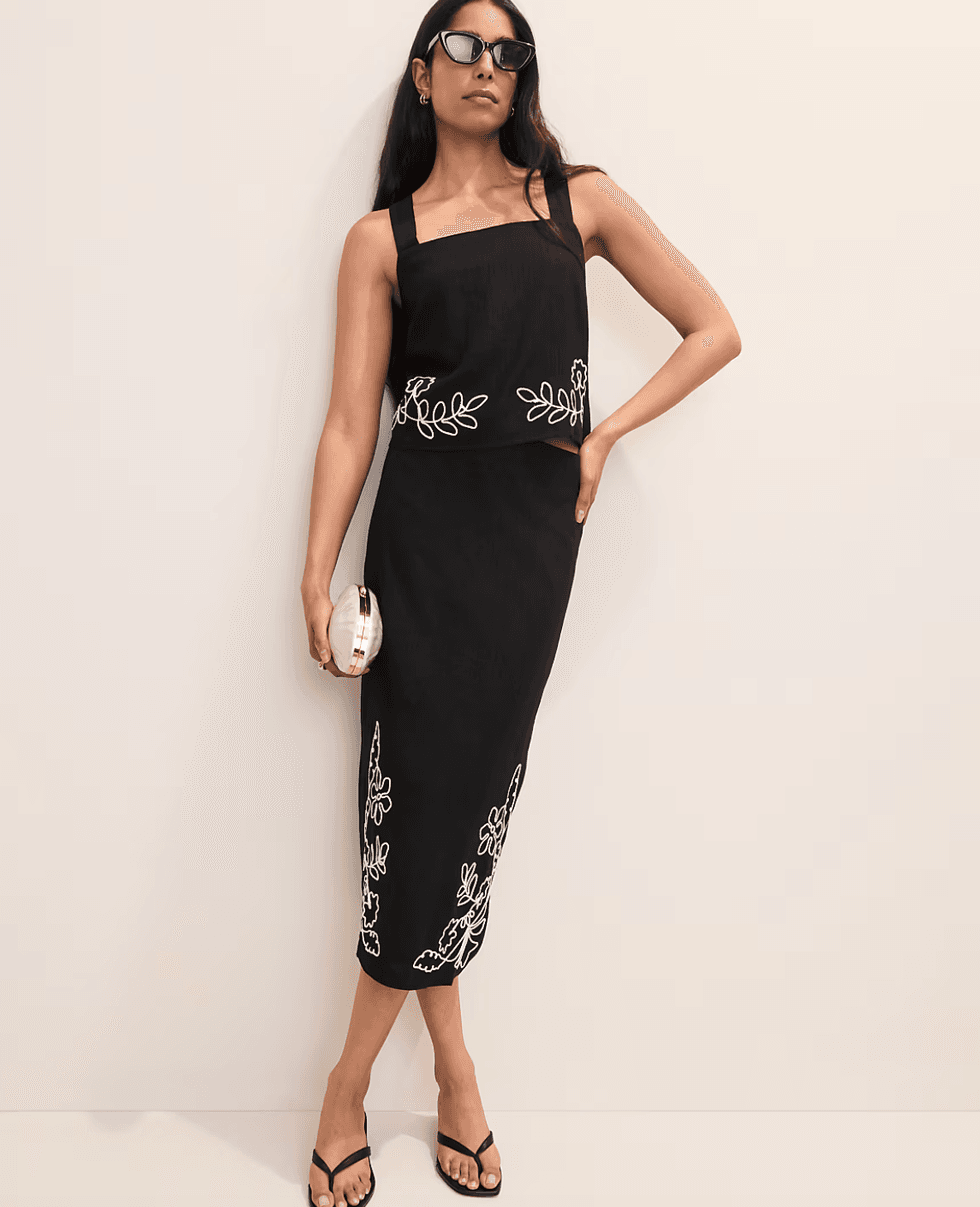 Woman in black floral dress, sunglasses, holding a small clutch.