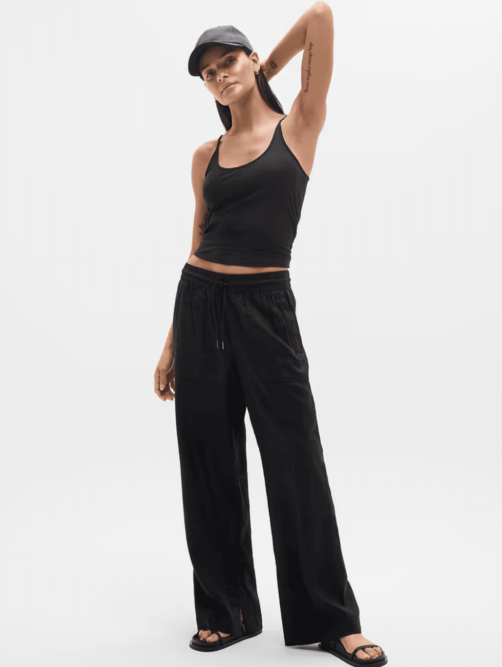Woman in black tank top and pants, wearing a cap, poses confidently against a white background.