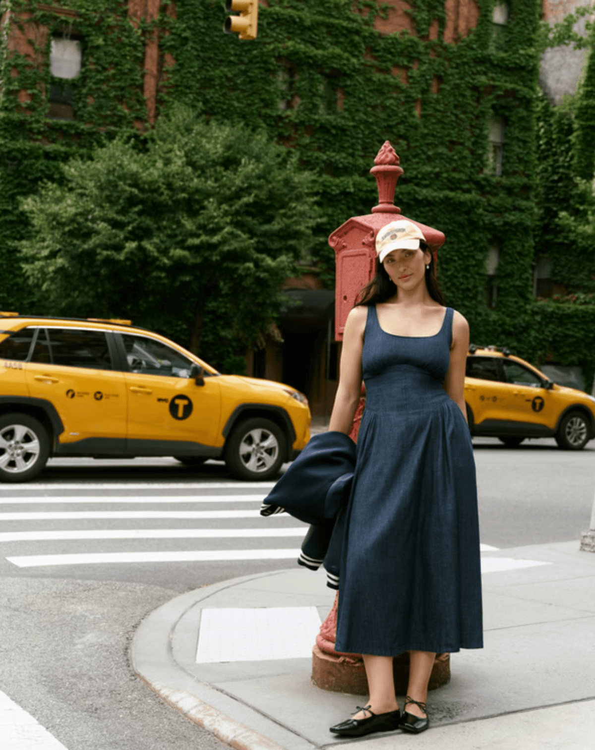 Woman in blue dress and cap stands by a red hydrant, with taxis and ivy-covered building.