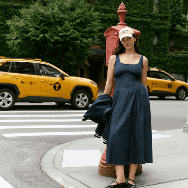 Woman in blue dress and cap stands by a red hydrant, with taxis and ivy-covered building.