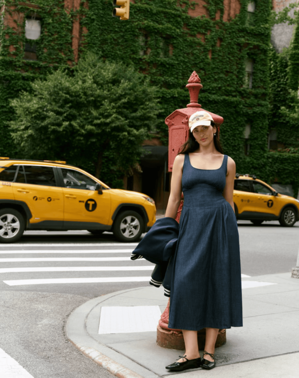 Woman in blue dress and cap stands by a red hydrant, with taxis and ivy-covered building.