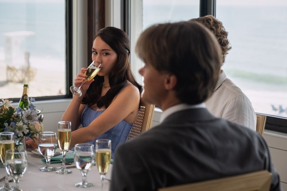 Woman in blue dress drinks champagne at a seaside dining table, surrounded by other guests lola tung as belly conklin