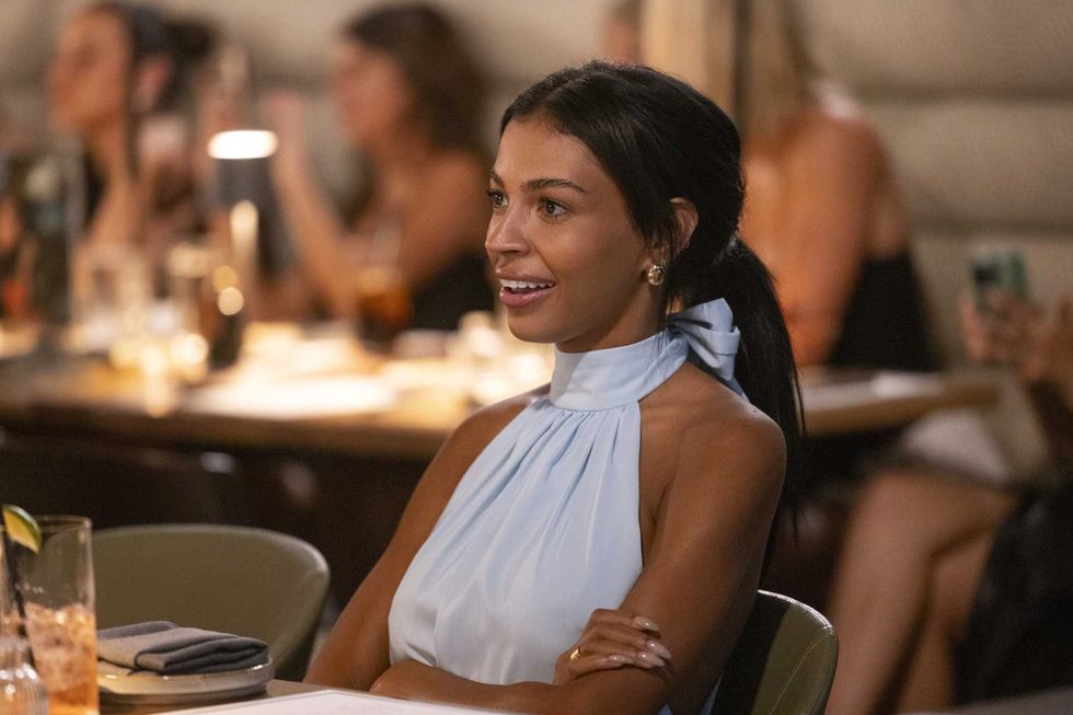 Woman in blue dress sitting at a restaurant table, smiling with arms crossed.