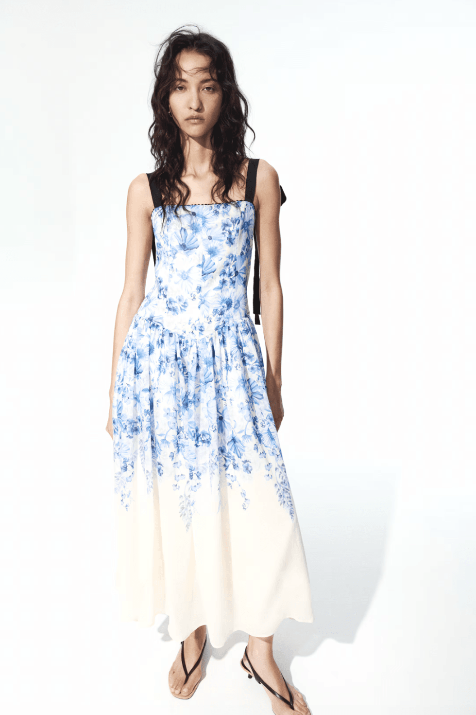 Woman in blue floral dress with black straps, standing on white background.