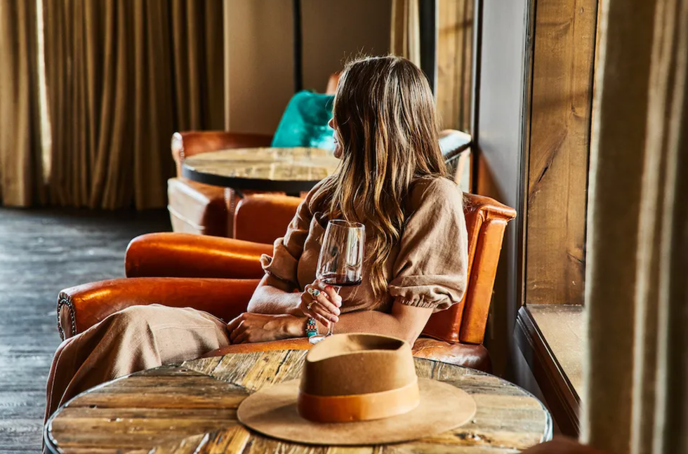 Woman in brown dress with wine glass, sitting on an orange armchair, looking out the window.