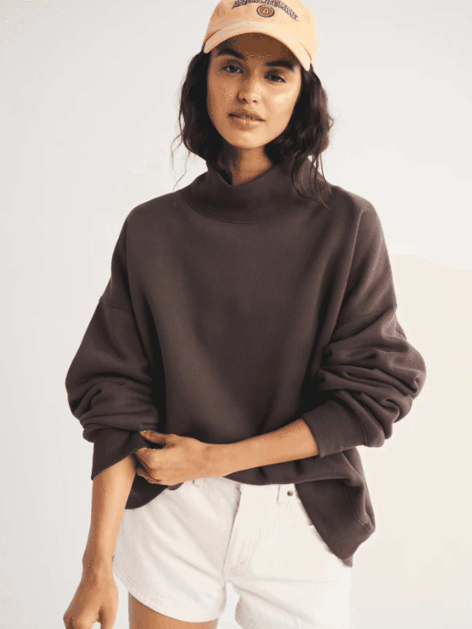 Woman in brown sweater, white shorts, and beige cap against plain background.
