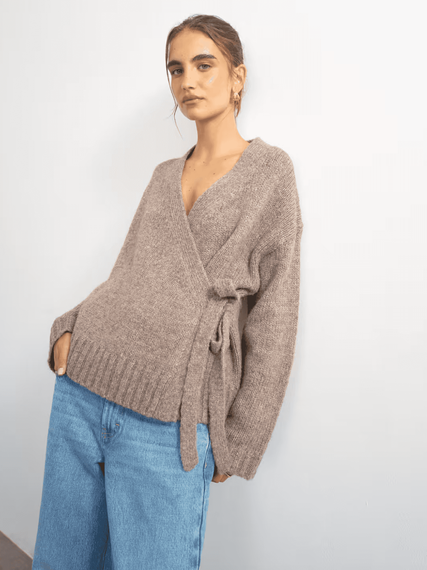 Woman in brown wrap sweater and blue jeans against white wall.