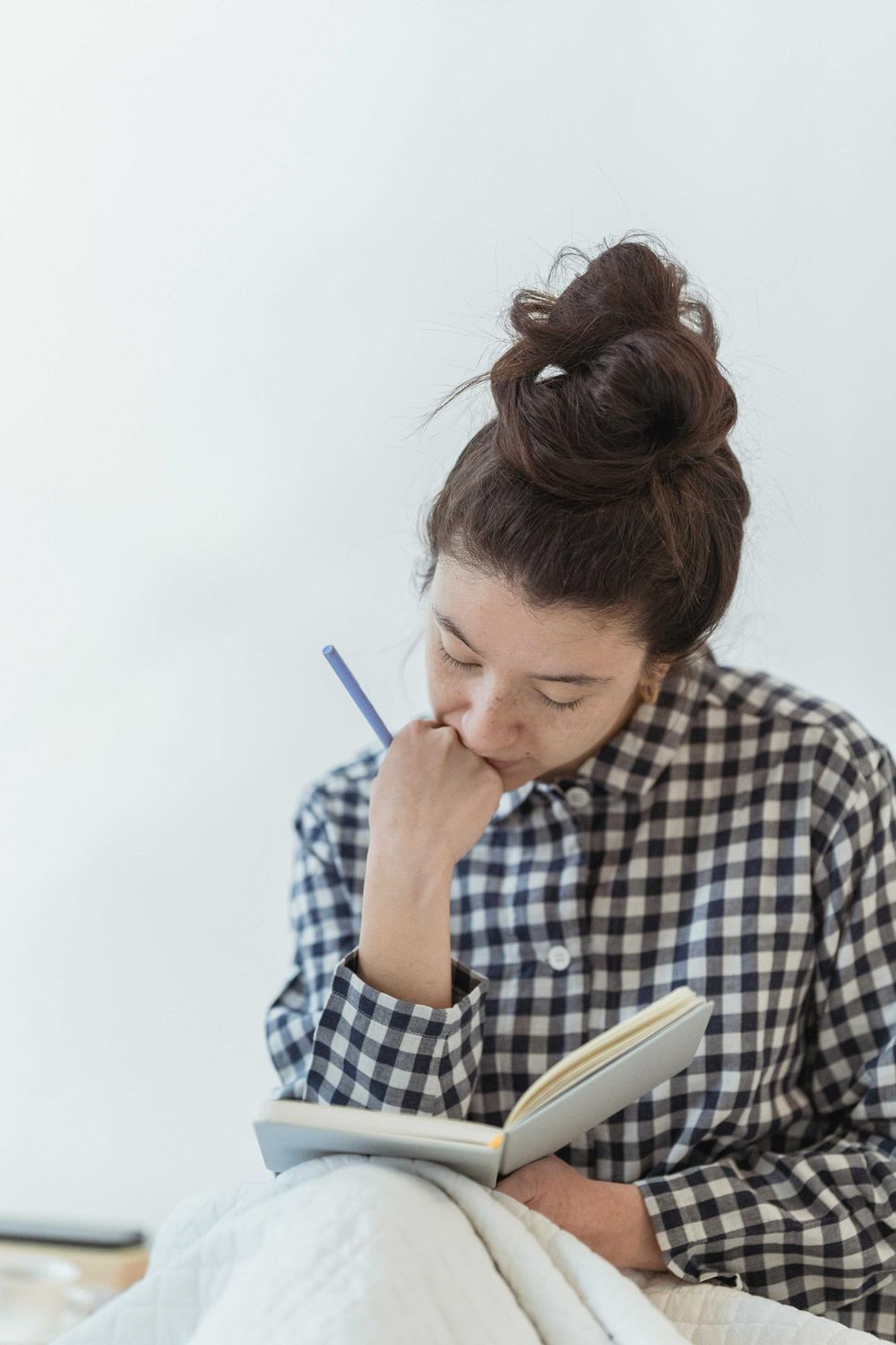 Woman in checkered shirt reading a book, holding a pen thoughtfully.