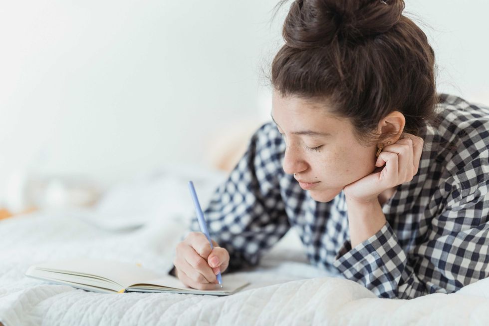 Woman in checkered shirt writing in a notebook on a bed.