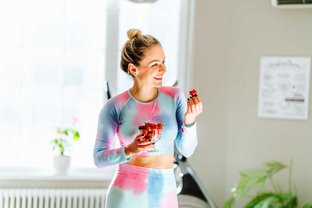 Woman in colorful outfit holds bowl of strawberries, standing in bright room with plants.