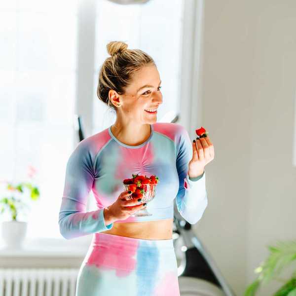 Woman in colorful outfit holds bowl of strawberries, standing in bright room with plants.