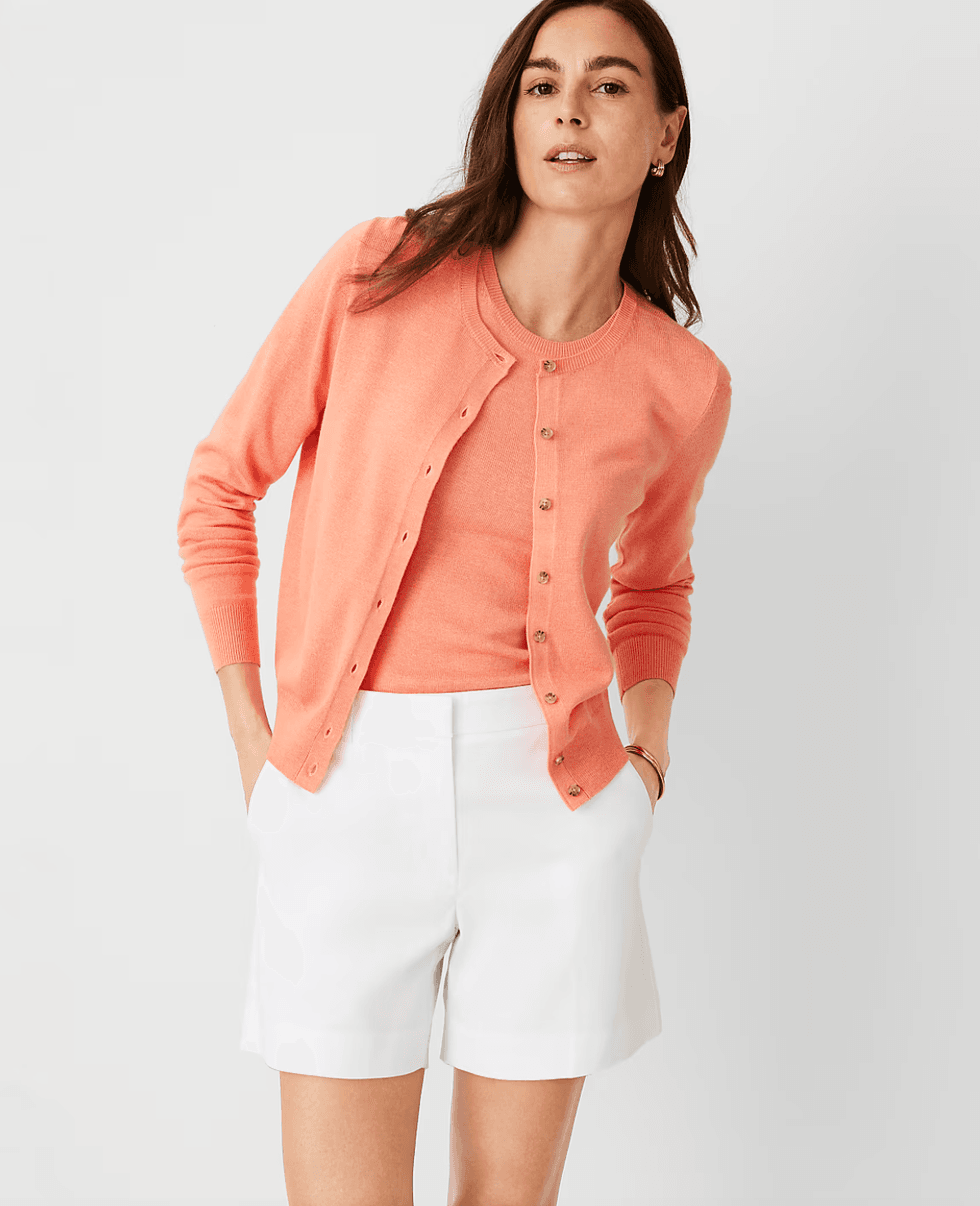 Woman in coral cardigan and top with white shorts, posing against a light background.