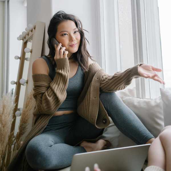 Woman in cozy attire talking on phone by a window, gesturing with one hand.