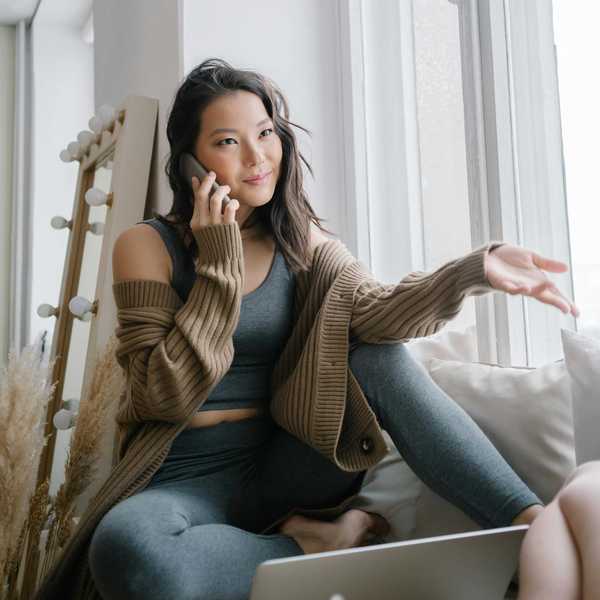 Woman in cozy attire talking on the phone while sitting by a window with a laptop.