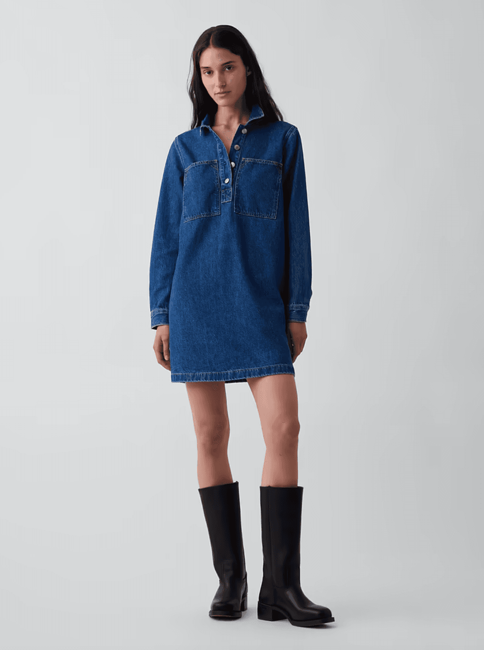 Woman in denim dress and black knee-high boots against a plain background.