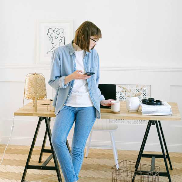 Woman in denim, holding phone and coffee, leans on table with art supplies and lamp.