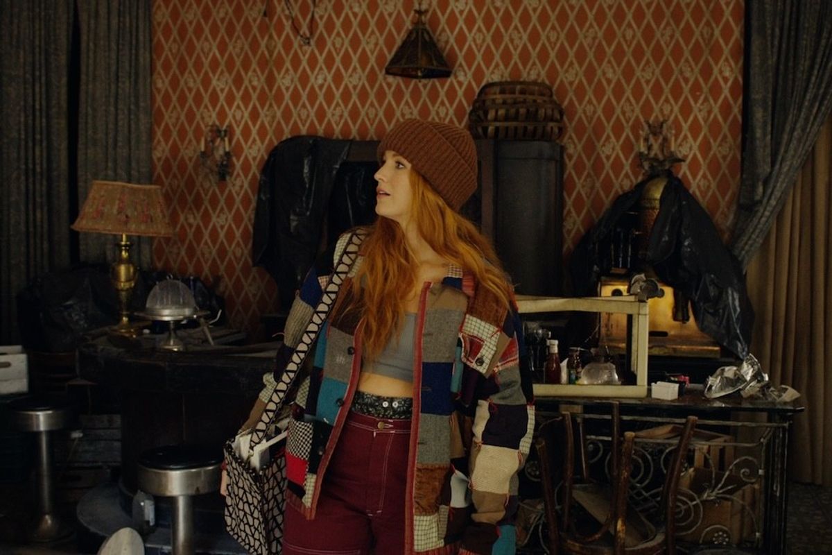 Woman in eclectic outfit stands in a cluttered, vintage-style room with patterned wallpaper.