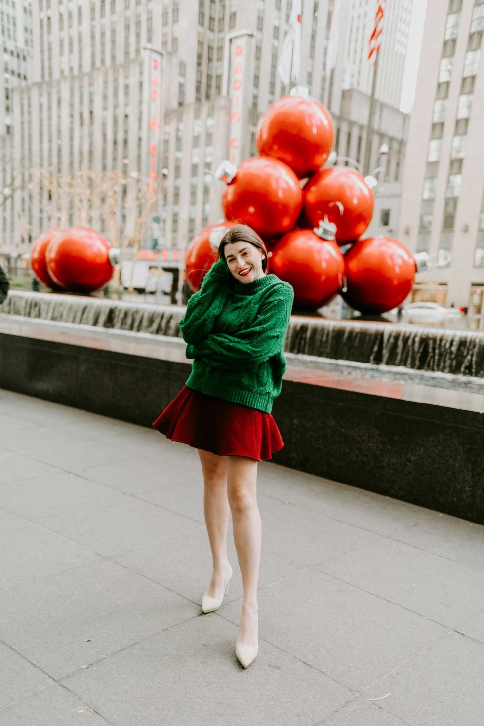 Woman in festive outfit smiles near giant red ornaments in a city plaza.