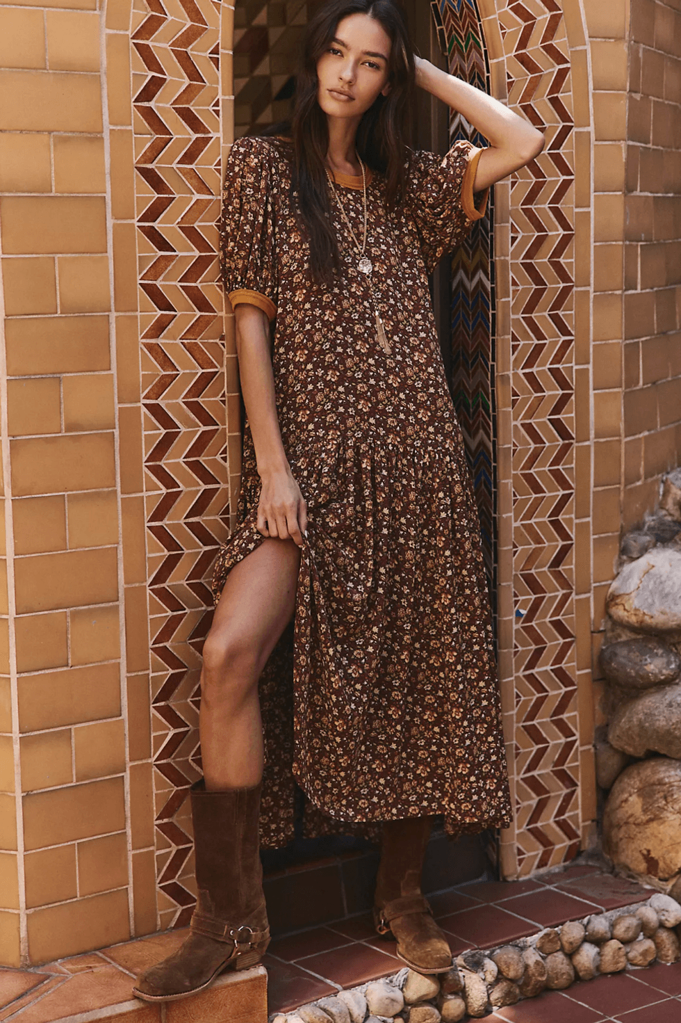 Woman in floral dress and boots poses by a tiled archway.