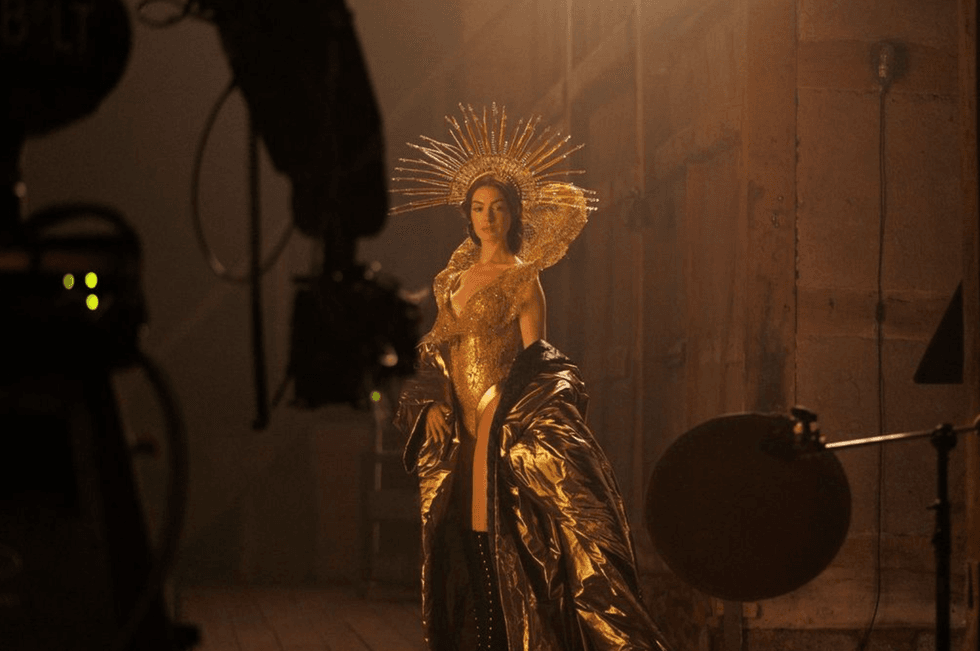 Woman in golden attire with a radiant headdress, standing in a dimly lit studio.