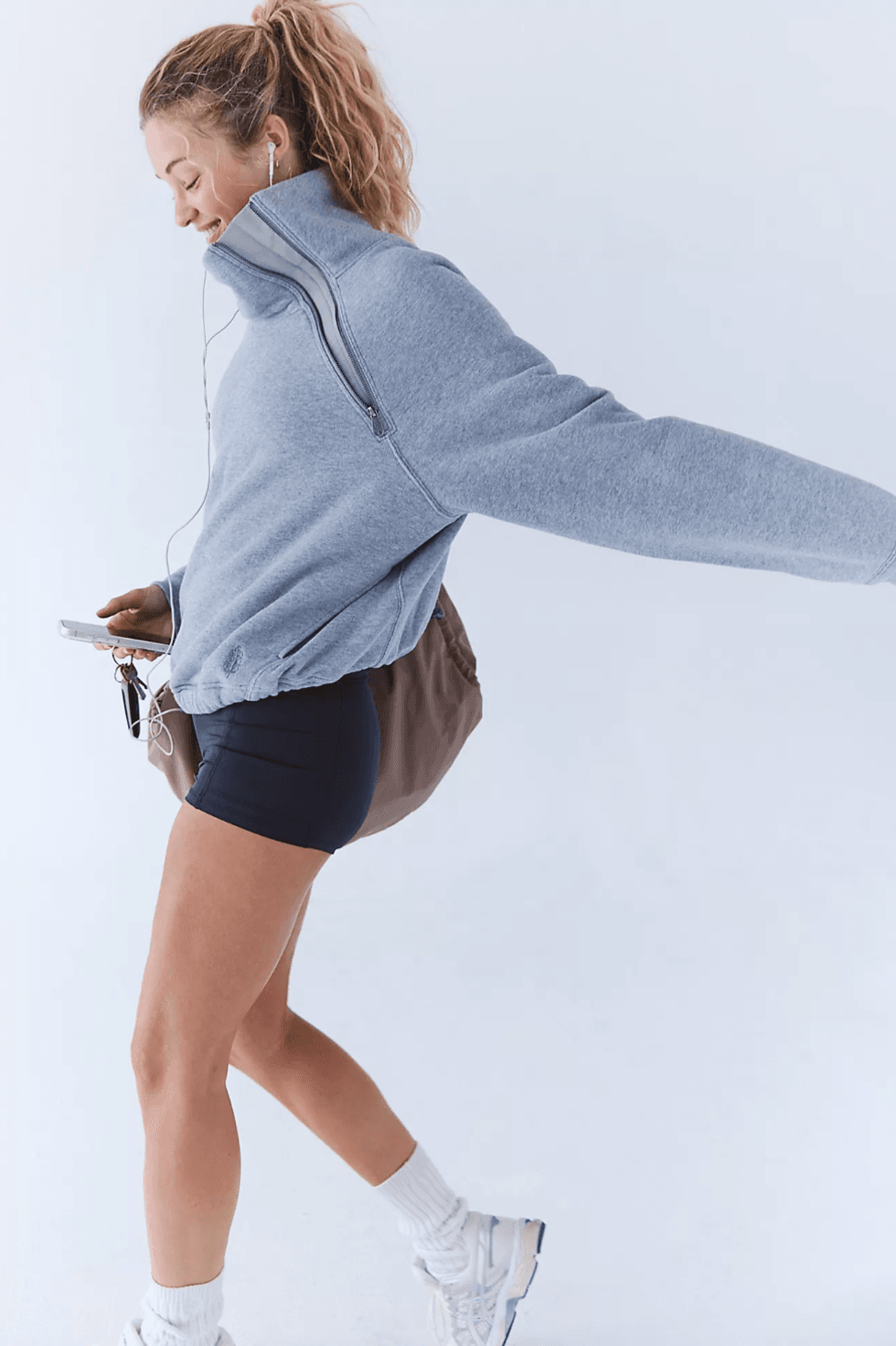 Woman in gray sweater walking, wearing earbuds and holding phone, against white background.