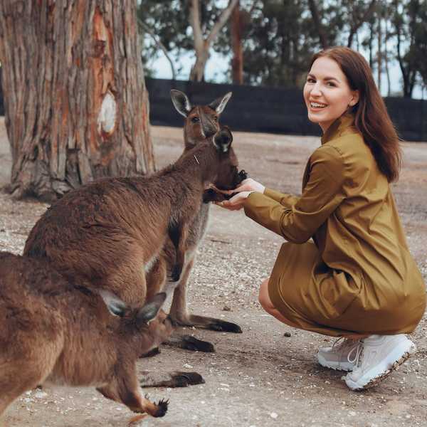Woman in green dress feeding kangaroos in a wooded area, smiling at the camera.