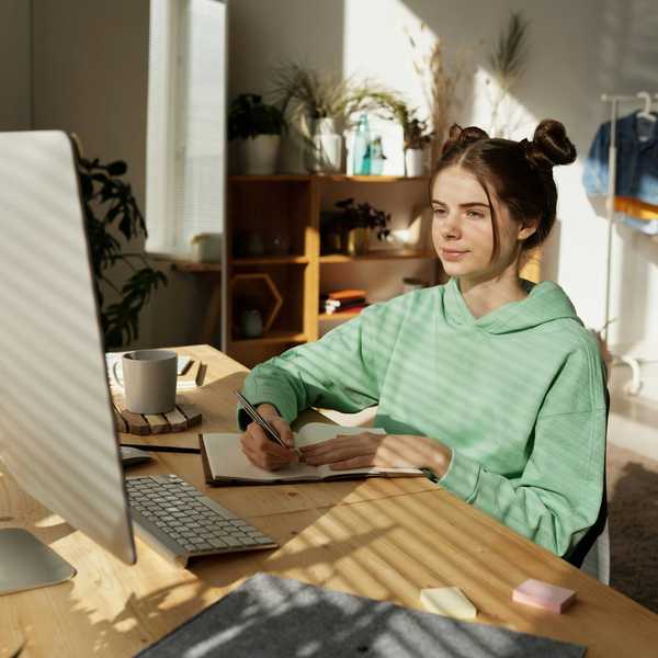 Woman in green hoodie working at a desk with a computer and notebook.