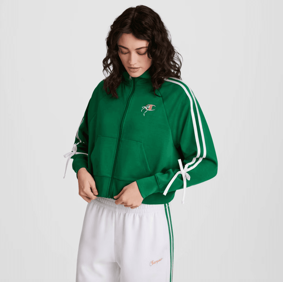 Woman in green track jacket and white pants, looking down.