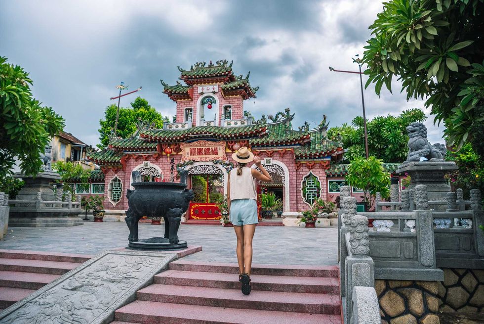 Woman in hat approaches ornate pink temple under cloudy sky.