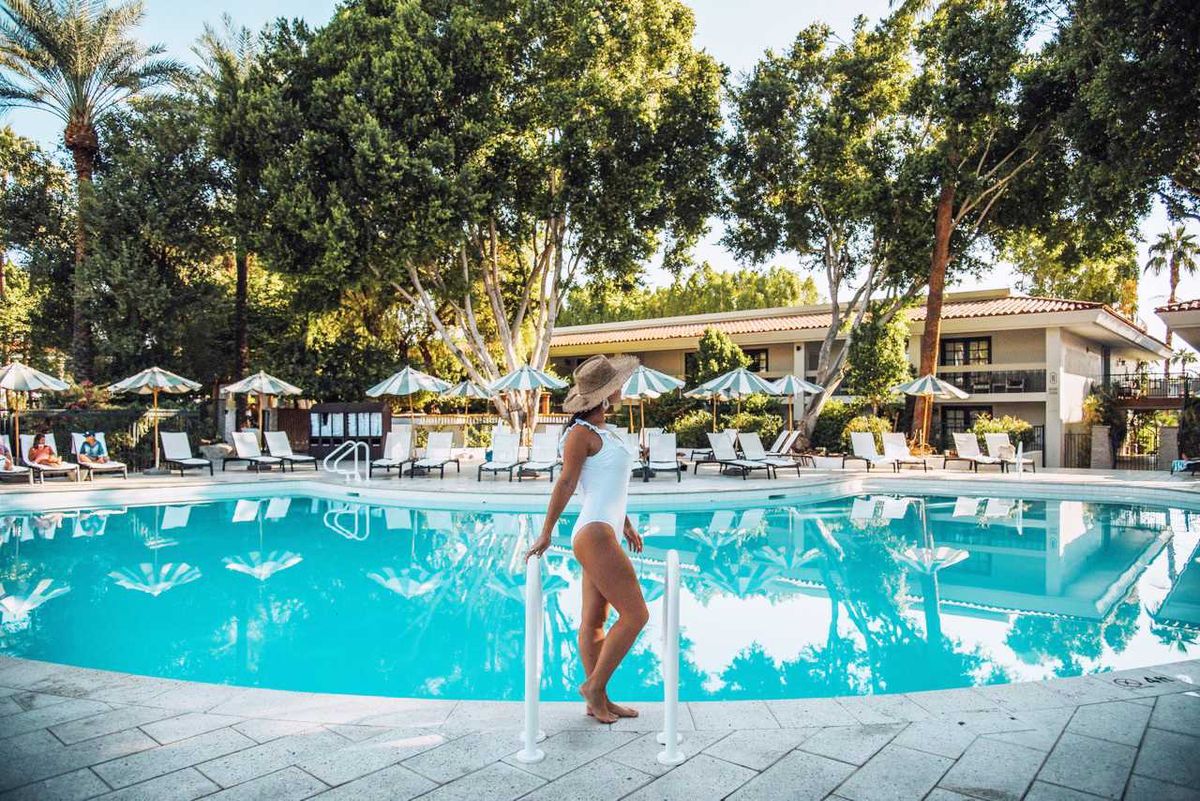 Woman in hat by a clear pool with lounge chairs and umbrellas under trees.