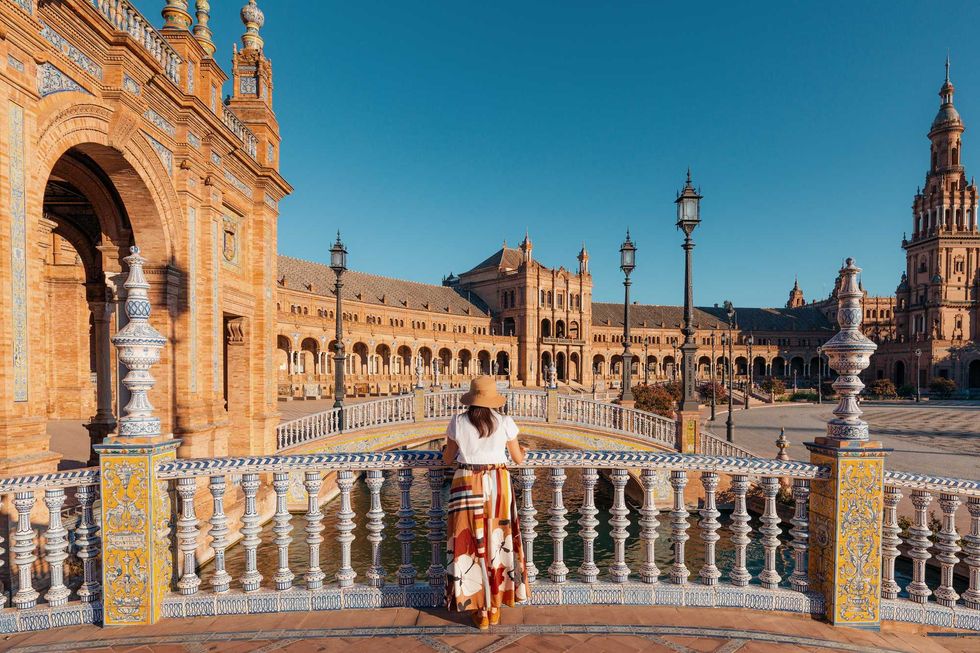Woman in hat overlooks ornate plaza with arches and towers in warm sunlight.