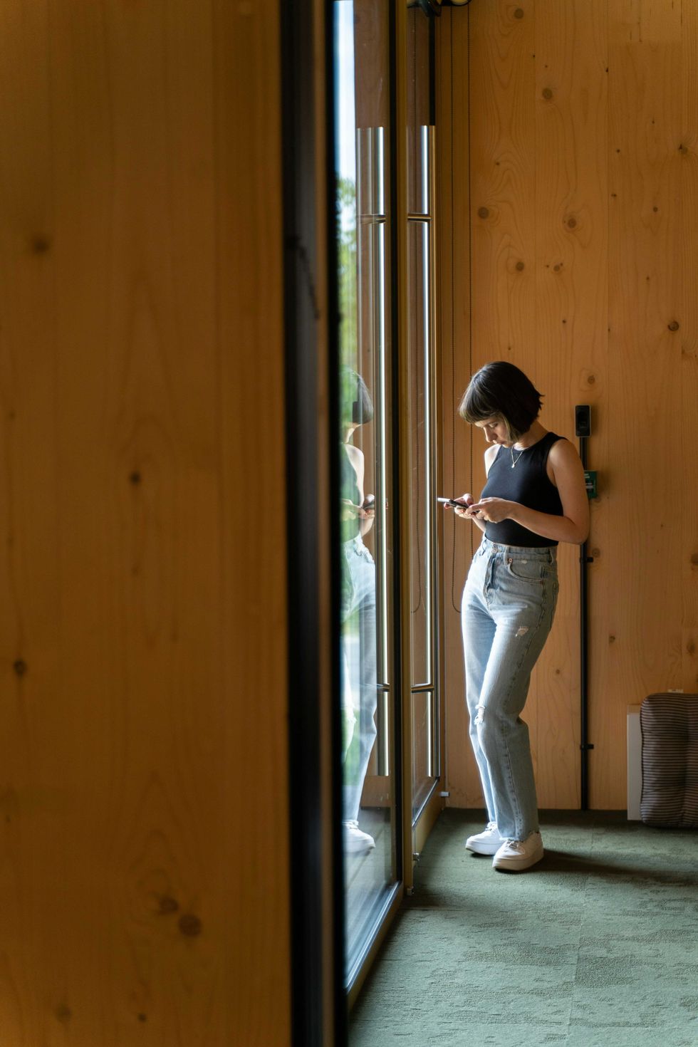 Woman in jeans and tank top, looking at phone by large window reflection.