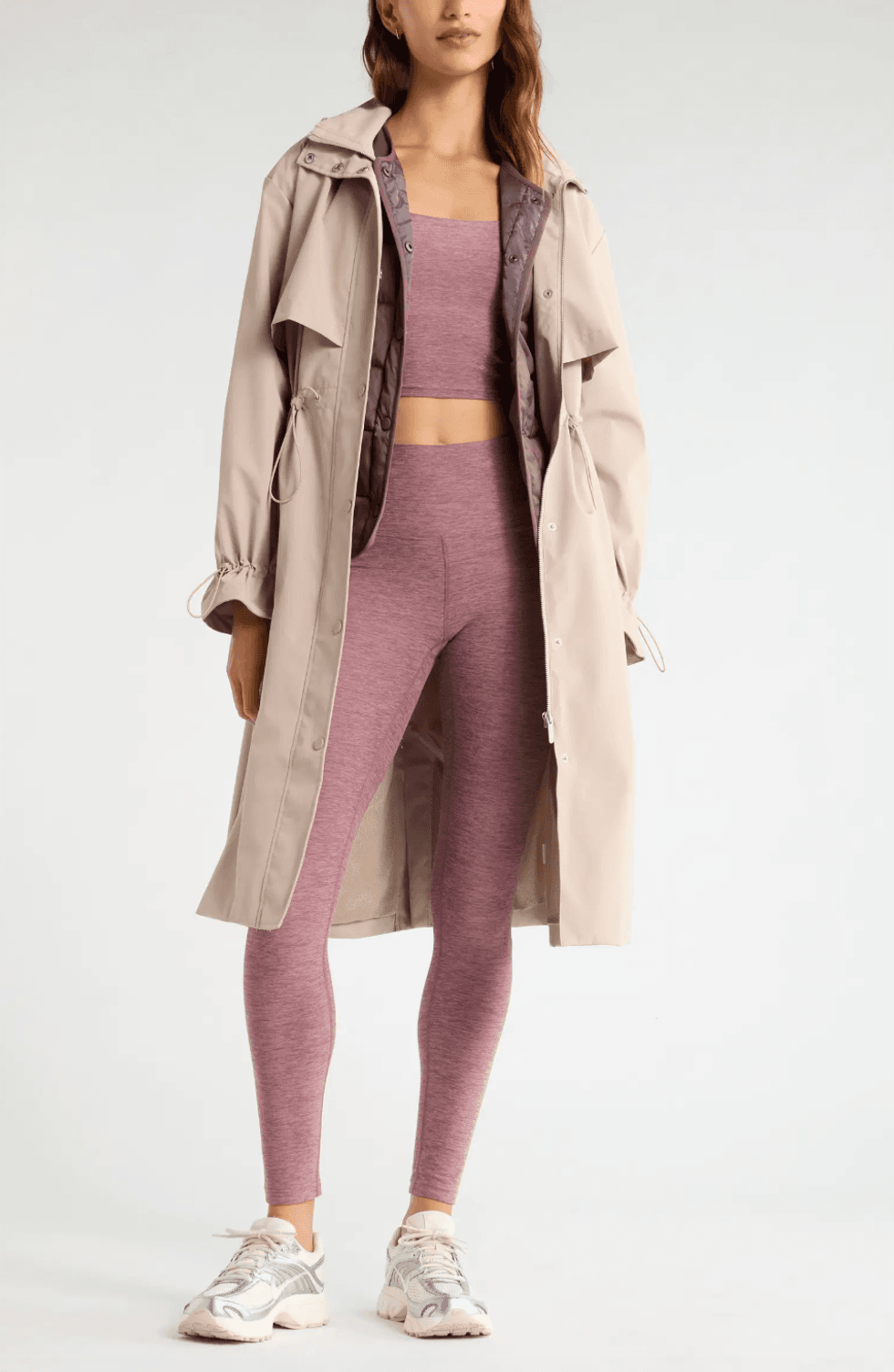 Woman in mauve activewear and beige coat with white sneakers.