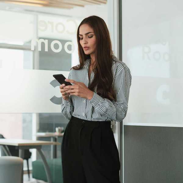 Woman in office attire using a smartphone in a modern meeting room.